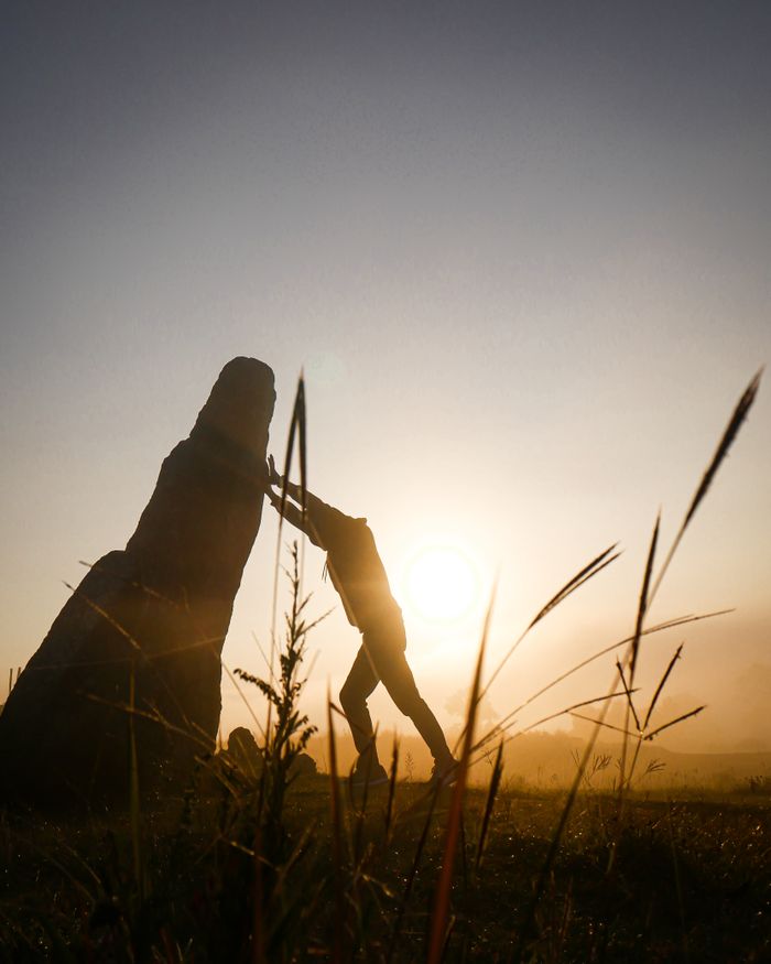 Dramatic silhouette of a person pushing a large stone in a field during sunset with warm tones.